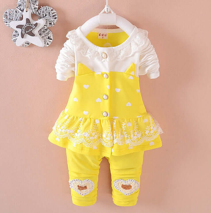 BibiCola Baby Girls- Yellow baby outfit with white ruffled top and heart details on a pink background