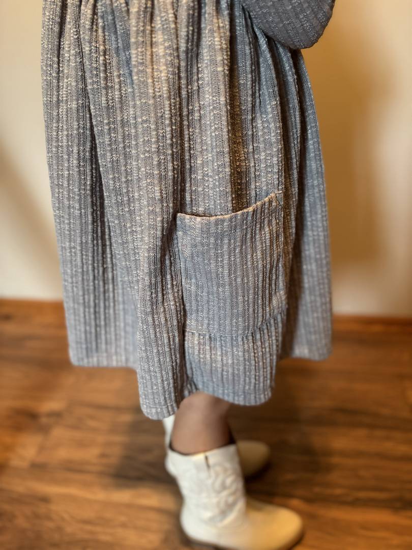 Person wearing a textured gray dress with a pocket on a wooden floor.