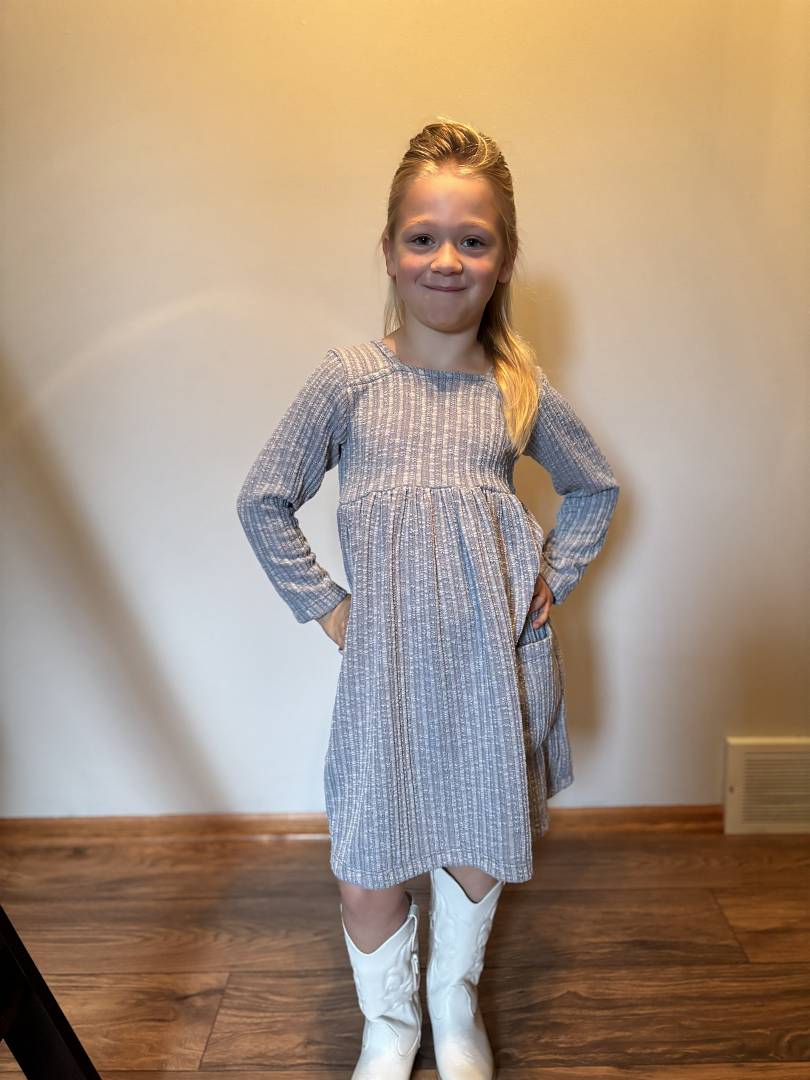 Young girl wearing a charcoal checkered dress and white boots standing against a plain wall.