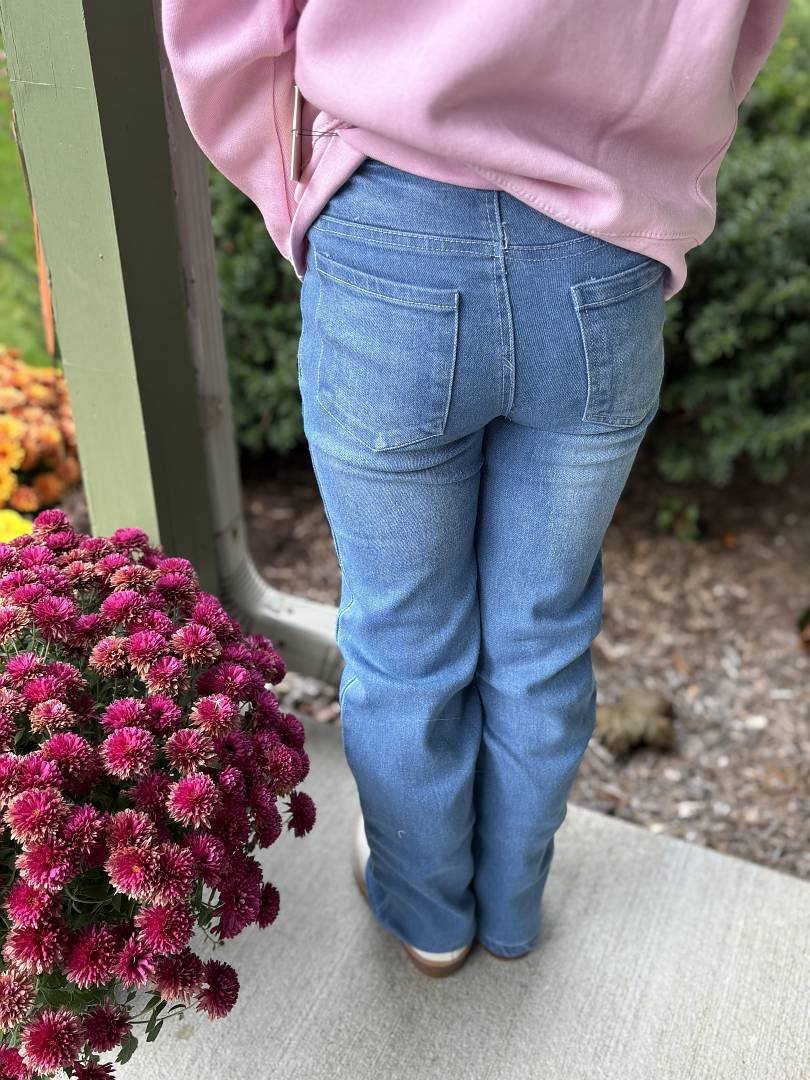 Thea Jeans - Person wearing a pink sweatshirt and blue jeans standing next to a potted plant with pink flowers.