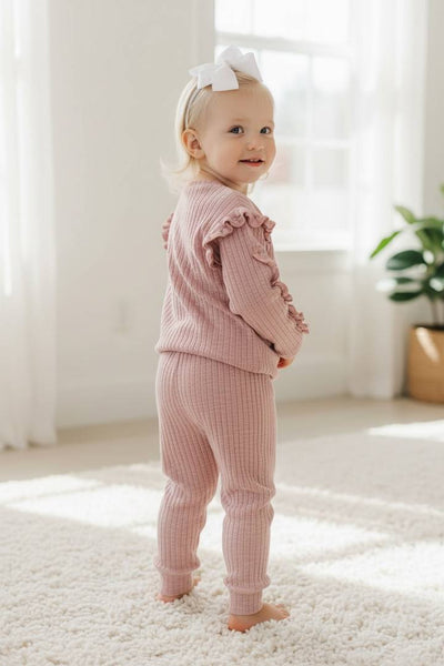 Baby in pink outfit standing on a light-colored floor with a white wall and plant in the background