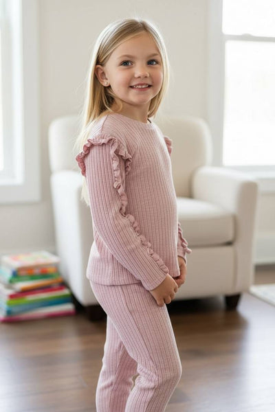 Young girl wearing a pink ribbed outfit with ruffled sleeves in a bright room.
