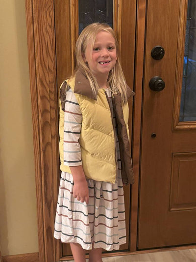 Young girl wearing a yellow vest and striped dress standing in front of a wooden door.