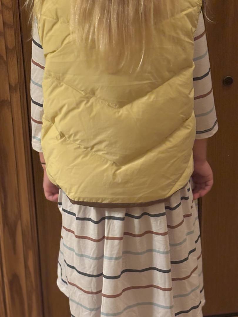 Person wearing a yellow puffer vest over a striped shirt and skirt against a wooden door background