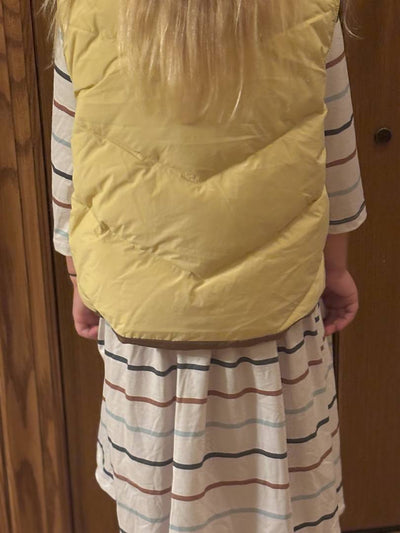 Person wearing a yellow puffer vest over a striped shirt and skirt against a wooden door background